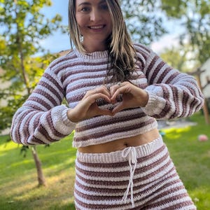 K&ouml;nnte beinhalten: Zweiteiliges H&auml;kelset in einem gestreiften Muster aus Wei&szlig; und Braun. Das Set beinhaltet einen lang&auml;rmligen, gecroppten Pullover und passende Shorts mit Kordelzug. Das Outfit wird im Freien bei nat&uuml;rlichem Licht gezeigt.