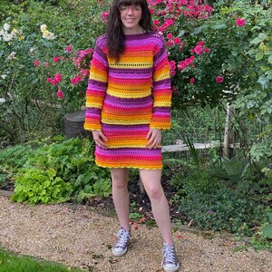 May include: A woman wearing a colourful striped crochet sweater and skirt. The sweater and skirt are made of a thick yarn and have a loose fit. The woman is standing in a garden with green grass and flowers.