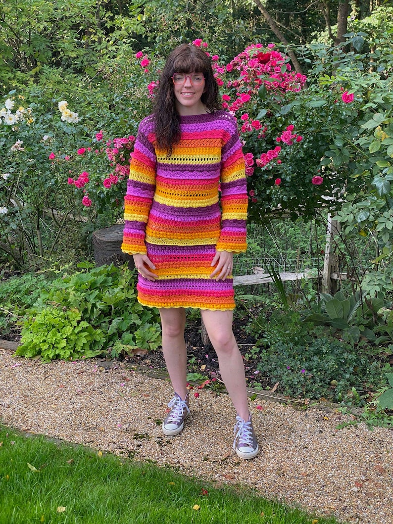 May include: A woman wearing a colourful striped crochet sweater and skirt. The sweater and skirt are made of a thick yarn and have a loose fit. The woman is standing in a garden with green grass and flowers.