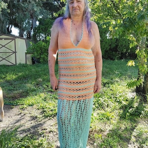 May include: A woman wearing a long, crocheted dress with a peach and teal color scheme. The dress has a V-neckline and a loose, flowing fit.