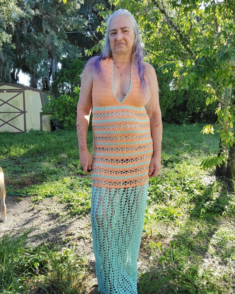 May include: A woman wearing a long, crocheted dress with a peach and teal color scheme. The dress has a V-neckline and a loose, flowing fit.