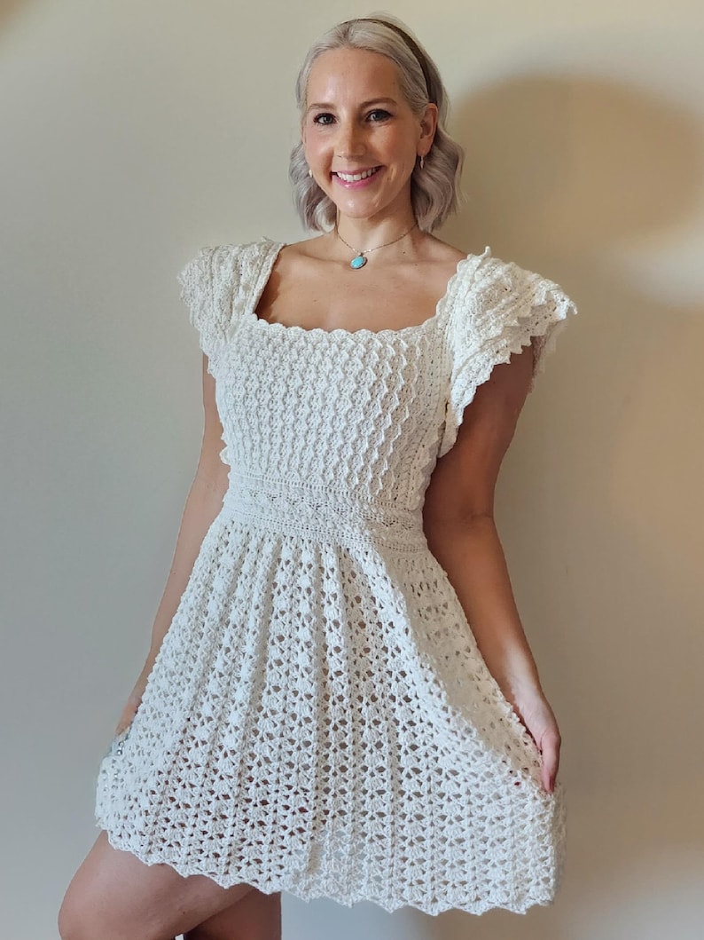 May include: A white crochet dress with a square neckline and short, ruffled sleeves. The dress has a fitted bodice and a flowing skirt.