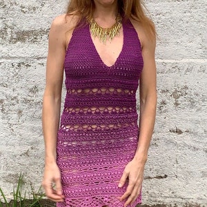 May include: A woman wearing a purple and pink crochet halter dress with a gold necklace. The dress has a gradient pattern and a crocheted design.