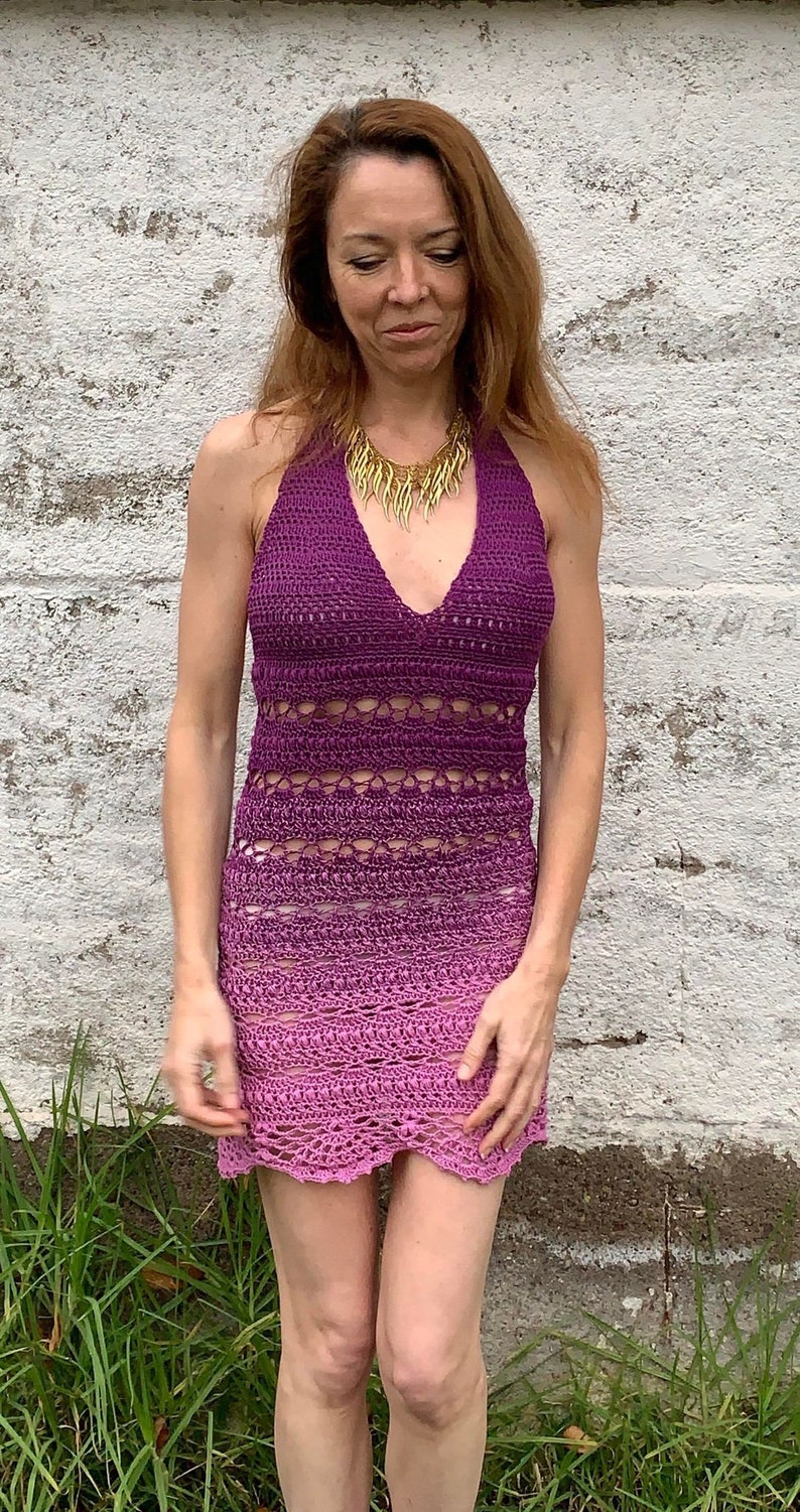 May include: A woman wearing a purple and pink crochet halter dress with a gold necklace. The dress has a gradient pattern and a crocheted design.
