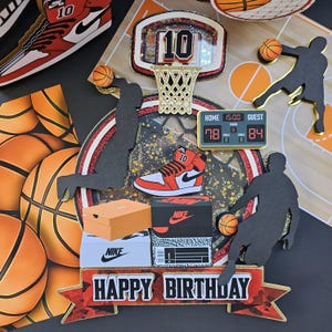May include: A red, white, and black basketball-themed cake topper with a basketball hoop, basketballs, and a pair of red and white sneakers. The cake topper also includes a scoreboard with the words "HOME" and "GUEST" and the numbers "78" and "84". The cake topper has a banner that says "HAPPY BIRTHDAY".