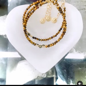 Könnte beinhalten: Drei übereinanderliegende Armbänder aus Tigerauge-Perlen und goldenen Akzenten. Die Armbänder werden auf einer weißen herzförmigen Oberfläche präsentiert. Die Bildunterschrift lautet "Bracelets"