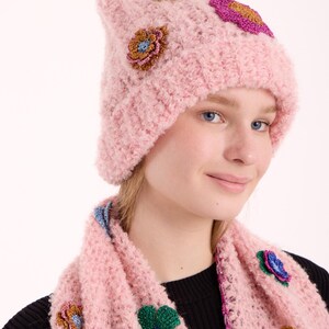 May include: A light pink knitted beanie and matching scarf set featuring textured fabric and decorative floral appliques in metallic gold, green, blue, and magenta. The set is crafted with a loose, open-knit pattern and a soft, fuzzy yarn texture.