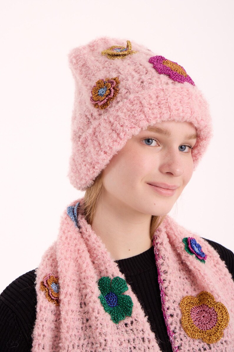 May include: A light pink knitted beanie and matching scarf set featuring textured fabric and decorative floral appliques in metallic gold, green, blue, and magenta. The set is crafted with a loose, open-knit pattern and a soft, fuzzy yarn texture.