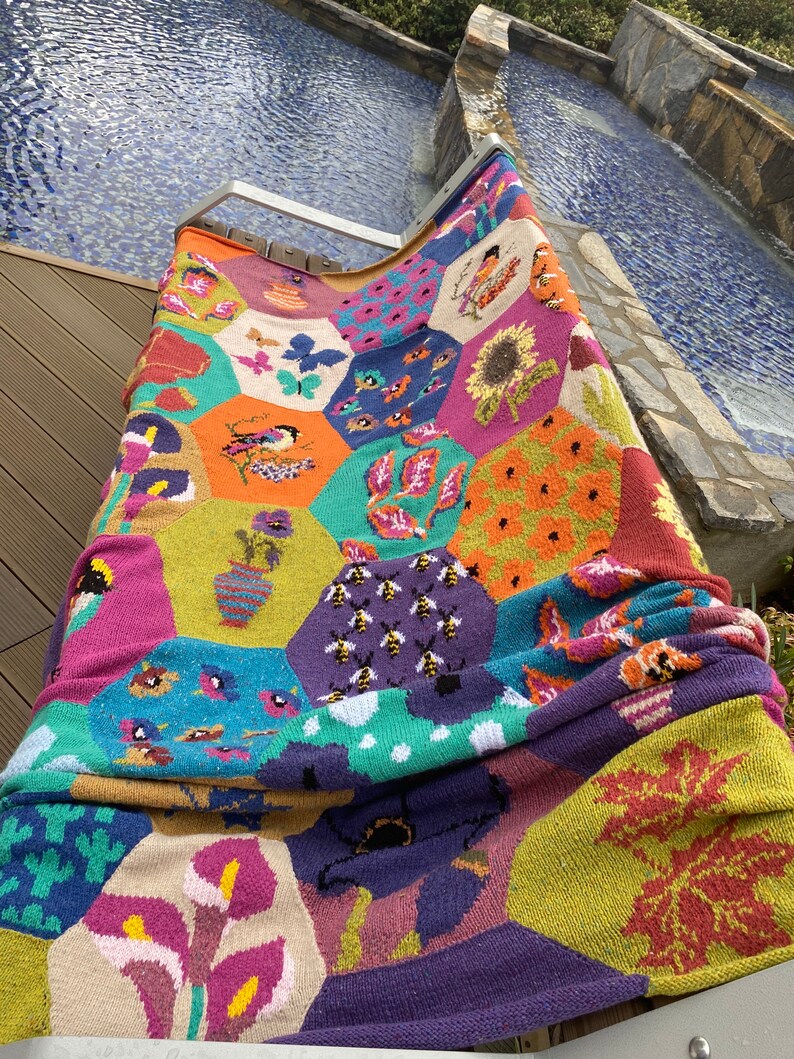 May include: A colorful knitted throw blanket with a hexagonal pattern featuring various nature-inspired designs, including bees, flowers, and leaves.