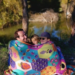 May include: A family of three is huddled together under a colorful patchwork blanket with a hexagonal pattern. The blanket features a variety of floral and geometric designs, including bees, flowers, and vases. The family is sitting outdoors in a grassy area near a body of water.