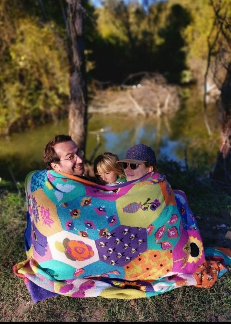 May include: A family of three is huddled together under a colorful patchwork blanket with a hexagonal pattern. The blanket features a variety of floral and geometric designs, including bees, flowers, and vases. The family is sitting outdoors in a grassy area near a body of water.