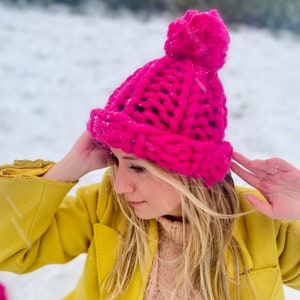 May include: A bright pink knitted beanie with a fluffy pom pom. The beanie is made with thick yarn and has a chunky knit texture.