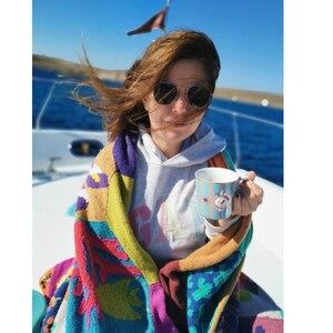 May include: A woman wearing sunglasses and a gray hoodie is wrapped in a colorful knitted blanket. She is holding a blue and white mug with a floral design.
