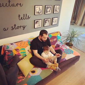 May include: A father and child sit on a couch reading a book. The couch has a colorful patchwork quilt. The wall behind them has a gallery of framed photos with the text "Every picture tells a story" written on it.