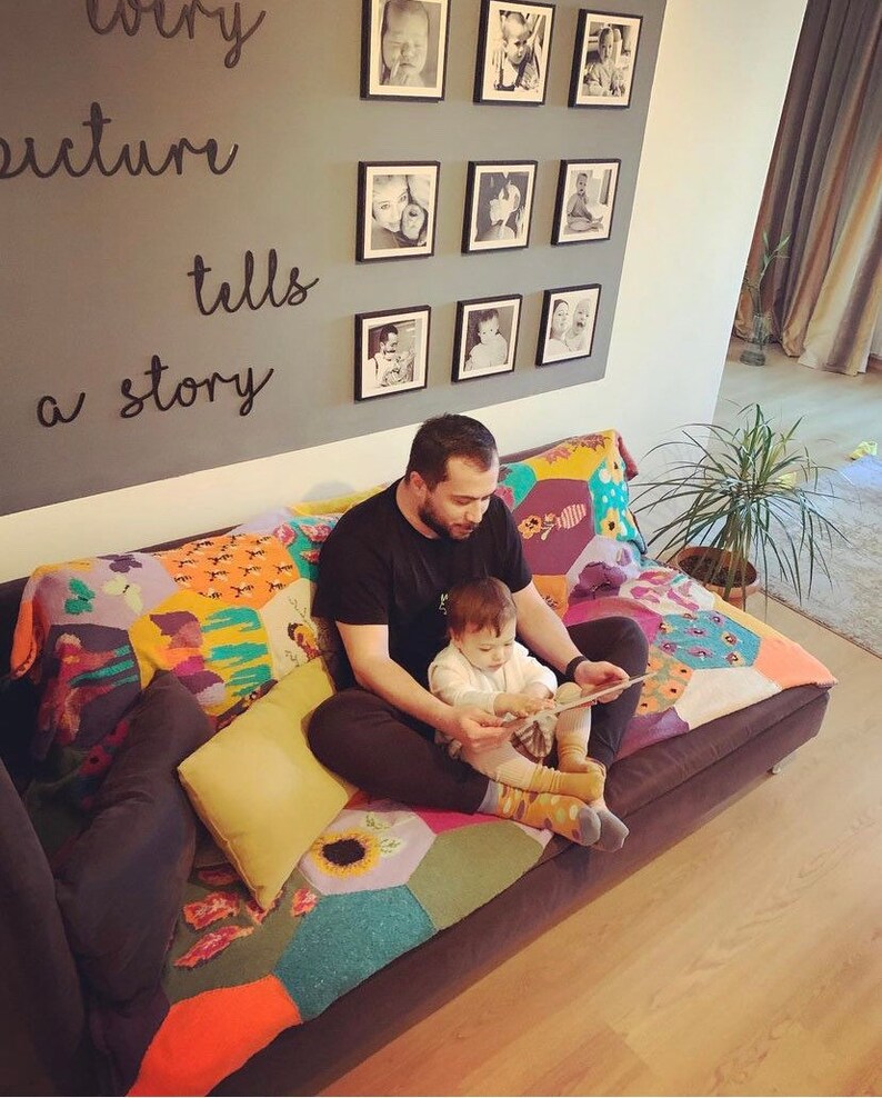 May include: A father and child sit on a couch reading a book. The couch has a colorful patchwork quilt. The wall behind them has a gallery of framed photos with the text "Every picture tells a story" written on it.