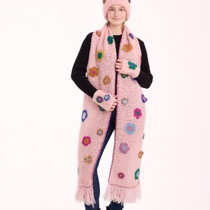 May include: A light pink crocheted set consisting of a beanie, a long scarf with fringe ends, and fingerless gloves. The set is decorated with various small, colorful crocheted flower appliqu&eacute;s in shades of blue, yellow, purple, and green.