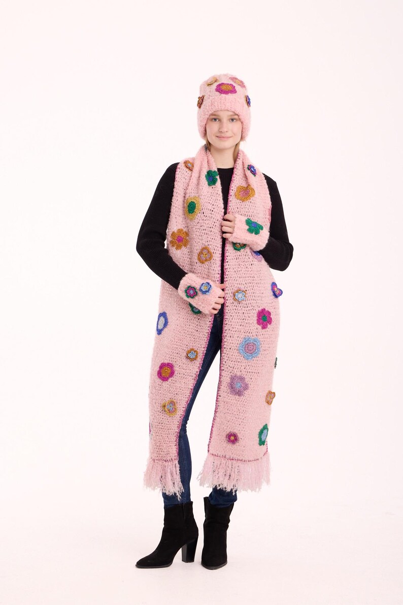 May include: A light pink crocheted set consisting of a beanie, a long scarf with fringe ends, and fingerless gloves. The set is decorated with various small, colorful crocheted flower appliqu&eacute;s in shades of blue, yellow, purple, and green.