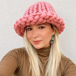 May include: A chunky knit bucket hat in a solid pastel pink color. The hat is crafted from thick, soft yarn with a prominent ribbed texture and a wide, downward-sloping brim that encircles the head, providing a thick and cozy appearance.