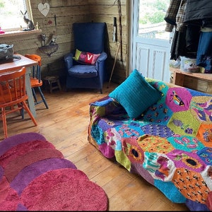 May include: A cozy living room with a colorful knitted throw blanket on a sofa, a blue armchair, a wooden table with orange chairs, and a rug with a multi-colored design.
