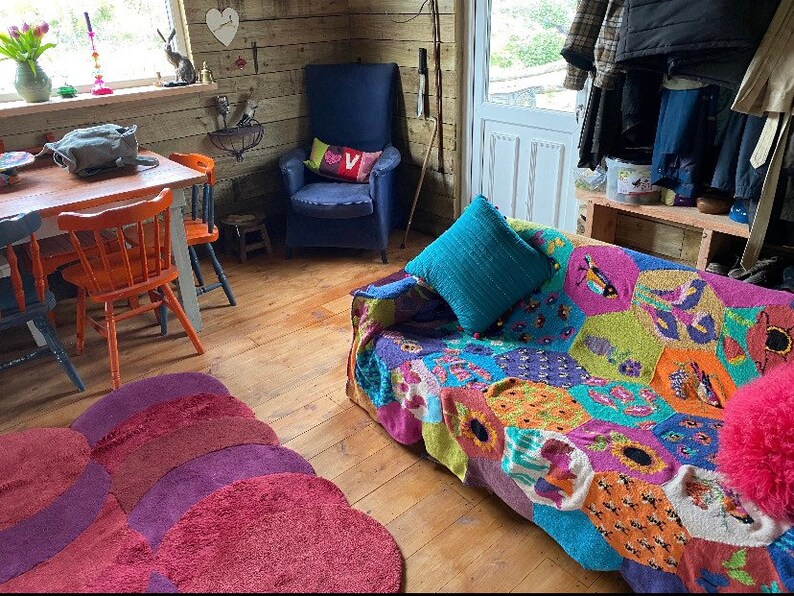 May include: A cozy living room with a colorful knitted throw blanket on a sofa, a blue armchair, a wooden table with orange chairs, and a rug with a multi-colored design.