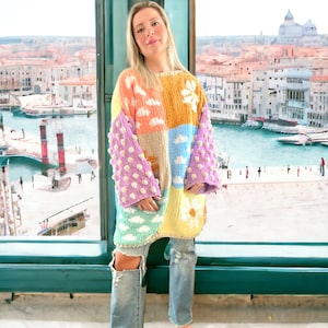 May include: A colorful patchwork sweater with a cloud and flower design. The sweater has puffy sleeves and is made of a soft, knitted fabric.