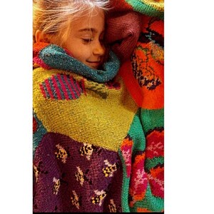 May include: A young person is wrapped in a colorful knitted blanket with a variety of patterns, including fish, bees, and flowers. The blanket is made of different colored yarns, including green, yellow, purple, orange, and red.