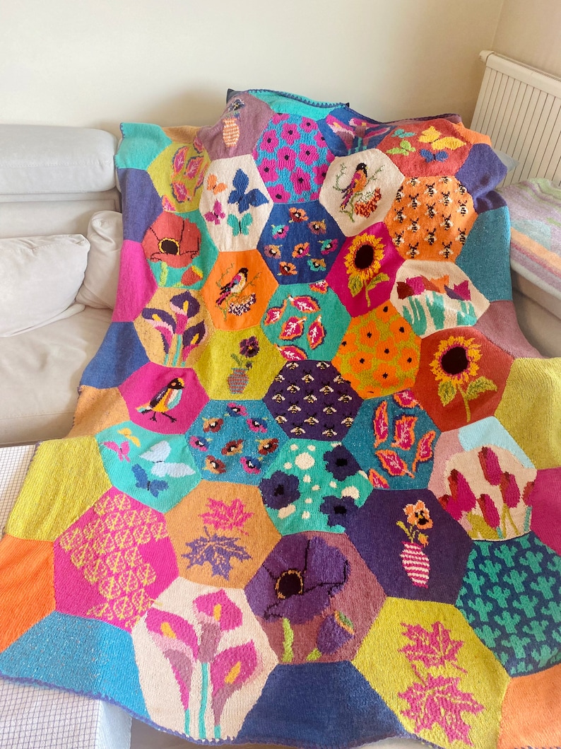 May include: A colorful knitted throw blanket with a hexagonal pattern featuring various floral and nature-inspired designs, including birds, flowers, leaves, and bees.