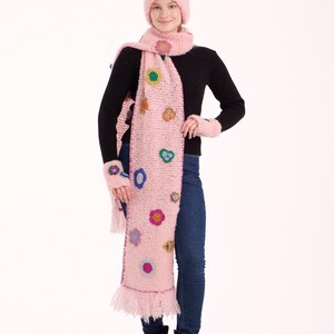 May include: A light pink knitted set consisting of a beanie, a long scarf with fringe, and fingerless gloves. Each piece is decorated with small, multi-colored crocheted flower appliqu&eacute;s in shades of yellow, blue, purple, and green.