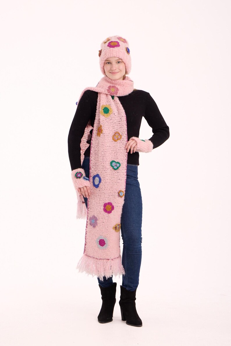 May include: A light pink knitted set consisting of a beanie, a long scarf with fringe, and fingerless gloves. Each piece is decorated with small, multi-colored crocheted flower appliqu&eacute;s in shades of yellow, blue, purple, and green.