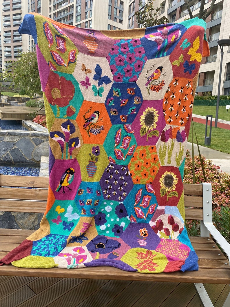 May include: A colorful knitted throw blanket with a hexagonal pattern featuring various floral and animal motifs, including butterflies, birds, and sunflowers. The blanket is made with a variety of yarn colors, including pink, blue, green, yellow, and orange.
