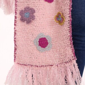 May include: A light pink crochet scarf featuring a fuzzy texture and a dark pink crocheted border. The scarf is decorated with several multi-colored floral appliques in shades of magenta, purple, silver, and gold, and finished with long fringe.