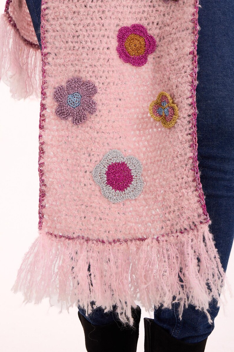 May include: A light pink crochet scarf featuring a fuzzy texture and a dark pink crocheted border. The scarf is decorated with several multi-colored floral appliques in shades of magenta, purple, silver, and gold, and finished with long fringe.
