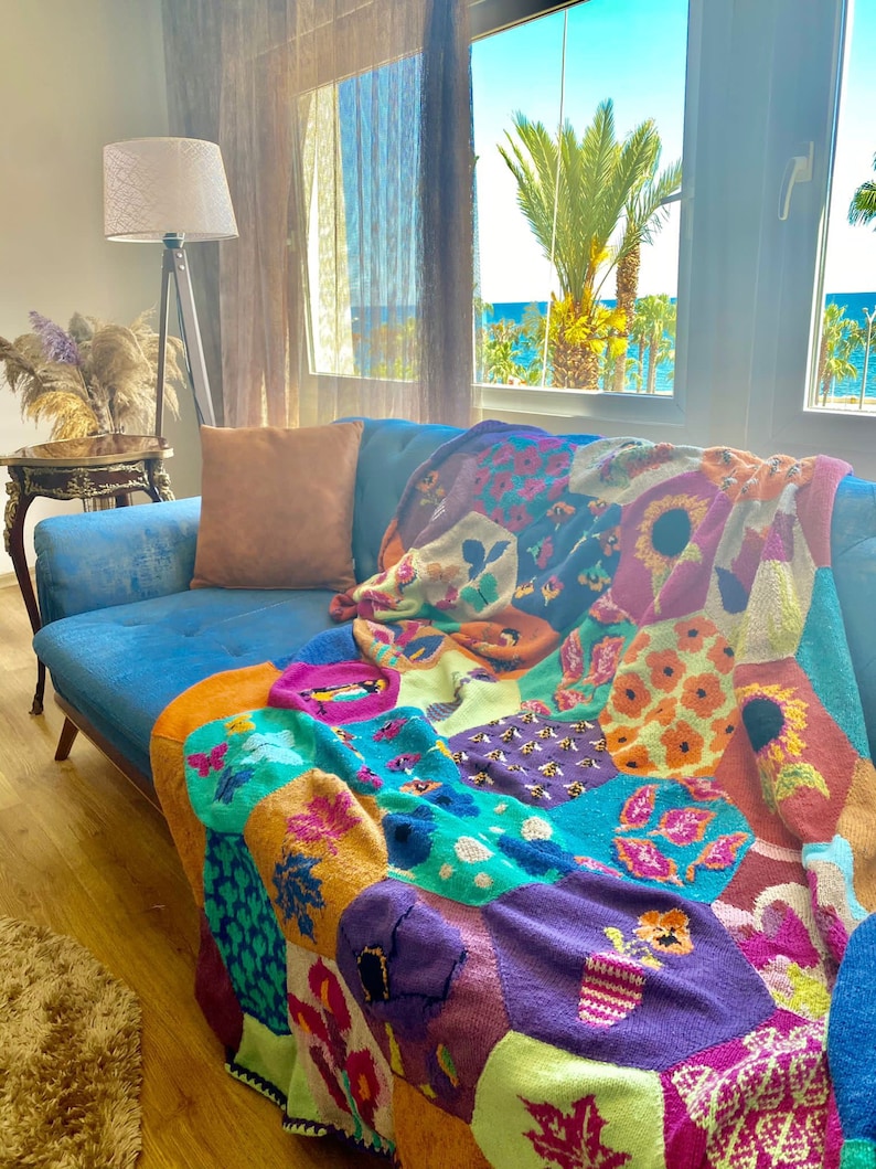 May include: A colorful knitted throw blanket with a patchwork design featuring various floral and geometric patterns in shades of orange, green, purple, and pink. The blanket is draped over a blue couch in a living room setting.