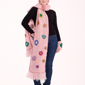 May include: A light pink crochet beanie, a long matching scarf with fringe ends, and fingerless gloves, all decorated with small, colorful crocheted flower appliqu&eacute;s in shades of blue, yellow, purple, and green, worn over a black sweater and jeans.