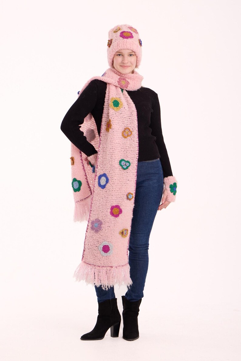 May include: A light pink crochet beanie, a long matching scarf with fringe ends, and fingerless gloves, all decorated with small, colorful crocheted flower appliqu&eacute;s in shades of blue, yellow, purple, and green, worn over a black sweater and jeans.