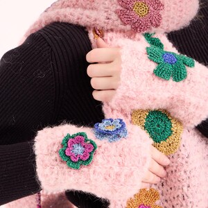 May include: A fuzzy light pink crocheted scarf and matching fingerless gloves. The set is decorated with various multi-colored crocheted flower appliqu&eacute;s in shades of green, gold, blue, and magenta, featuring a textured, metallic-like yarn finish.