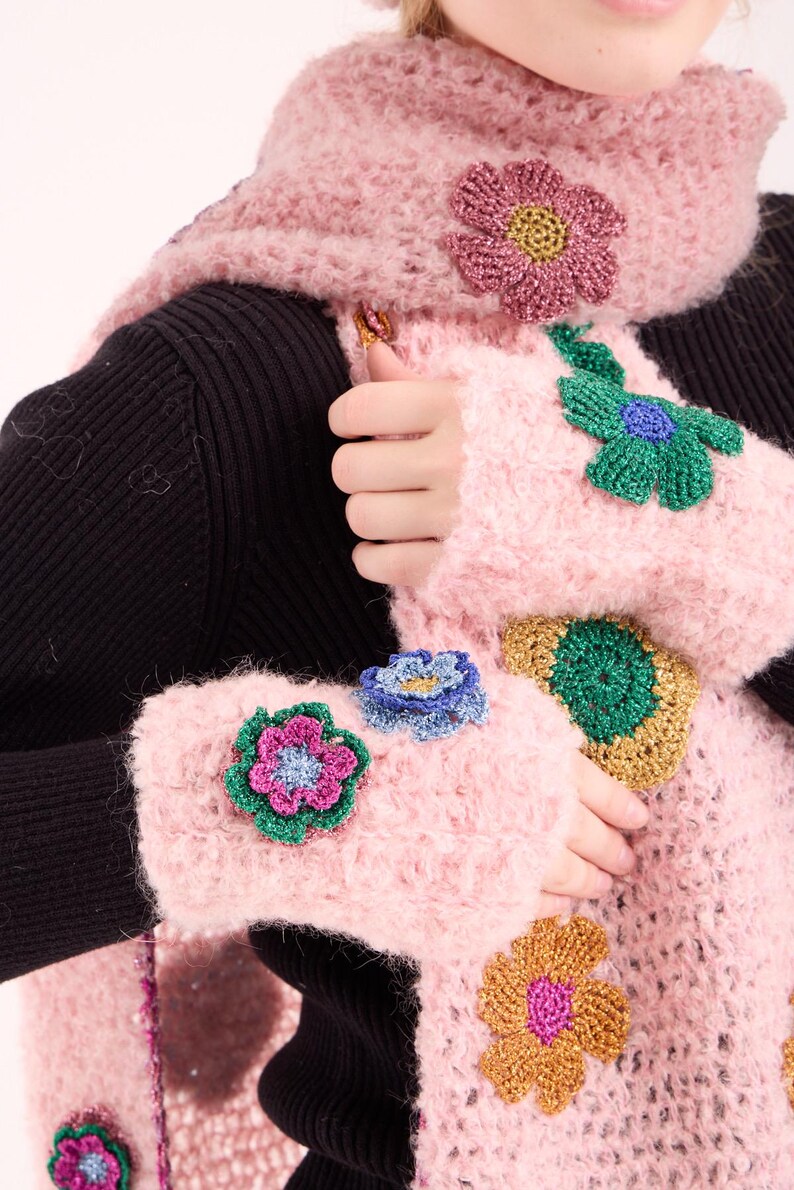 May include: A fuzzy light pink crocheted scarf and matching fingerless gloves. The set is decorated with various multi-colored crocheted flower appliqu&eacute;s in shades of green, gold, blue, and magenta, featuring a textured, metallic-like yarn finish.