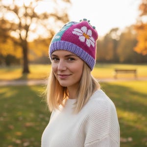 Può includere: Un berretto lavorato a maglia colorato con un motivo a margherita. Il cappello presenta sezioni rosa, verde acqua e viola, con una margherita bianca sul davanti. L'orlo è viola e la parte superiore ha piccoli pompon verdi. Il cappello è indossato da una persona con i capelli biondi.