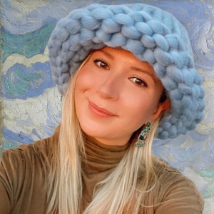 May include: A light blue, chunky knit hat with a wide brim. The hat is made of a soft, textured yarn and has a rounded crown. The woman is wearing a brown turtleneck and green earrings.