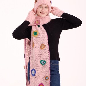 May include: A light pink fuzzy knit beanie, a matching long scarf, and fingerless gloves. The set features scattered, multi-colored crocheted flower appliqu&eacute;s in shades of yellow, blue, purple, and green, with a dark pink border on the scarf.