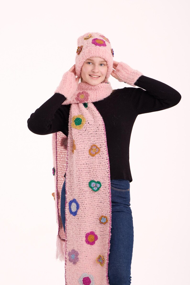 May include: A light pink fuzzy knit beanie, a matching long scarf, and fingerless gloves. The set features scattered, multi-colored crocheted flower appliqu&eacute;s in shades of yellow, blue, purple, and green, with a dark pink border on the scarf.