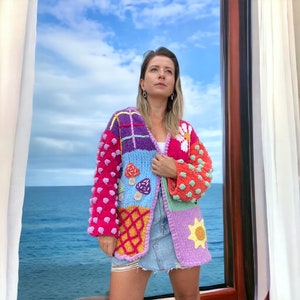 May include: A colorful crocheted cardigan with a patchwork design featuring flowers, mushrooms, and geometric patterns. The cardigan has a loose fit and long sleeves.