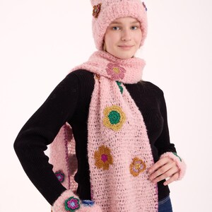 May include: A light pink fuzzy knit beanie, a matching long scarf, and fingerless gloves. Each piece features colorful crocheted flower appliqu&eacute;s in shades of blue, green, yellow, and purple with metallic thread accents throughout the set.