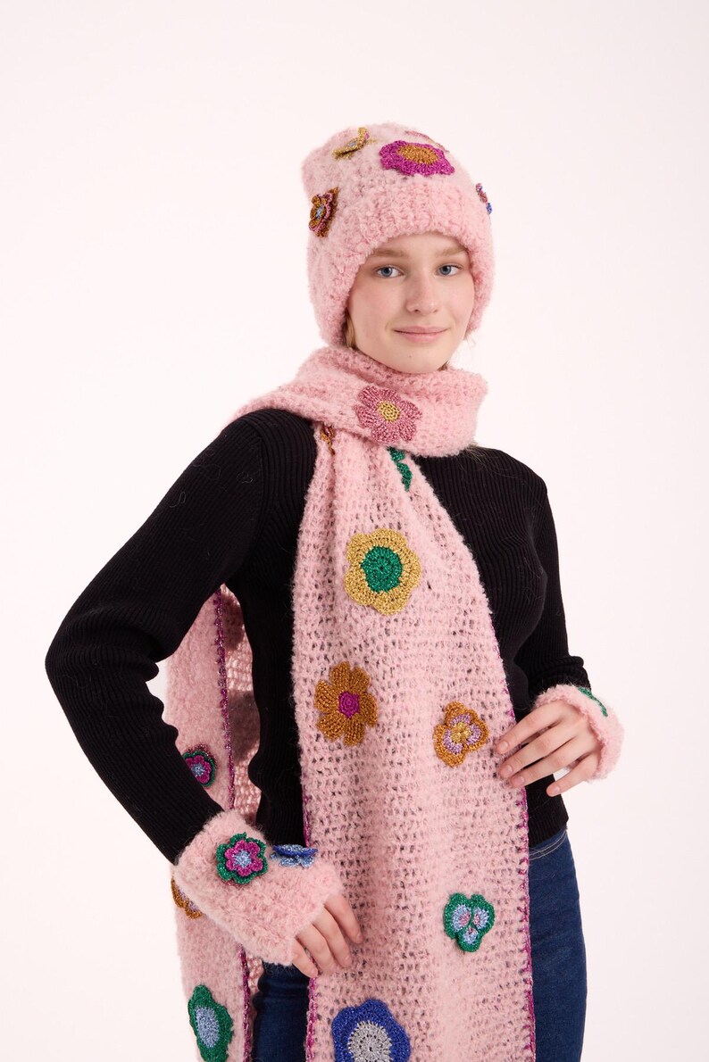 May include: A light pink fuzzy knit beanie, a matching long scarf, and fingerless gloves. Each piece features colorful crocheted flower appliqu&eacute;s in shades of blue, green, yellow, and purple with metallic thread accents throughout the set.