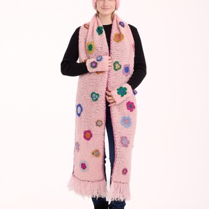 May include: A light pink crochet set consisting of a beanie, a long scarf with fringe ends, and fingerless gloves. The set is decorated with various small, multi-colored floral appliques in shades of blue, green, yellow, and purple.