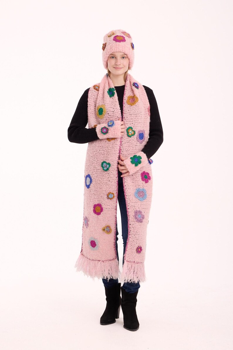 May include: A light pink crochet set consisting of a beanie, a long scarf with fringe ends, and fingerless gloves. The set is decorated with various small, multi-colored floral appliques in shades of blue, green, yellow, and purple.