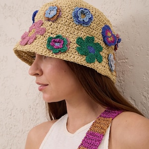 May include: A beige crocheted bucket hat adorned with multiple colorful, three-dimensional floral appliqués in shades of pink, blue, green, and purple. The hat features a textured, woven construction with a wide brim and a flat crown.