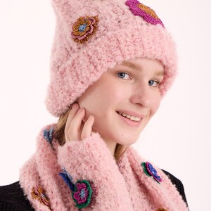 May include: A light pink knitted beanie and matching scarf set featuring a textured, fuzzy yarn construction. The set is decorated with multiple small, colorful crocheted flower appliqu&eacute;s in shades of gold, blue, purple, and magenta.