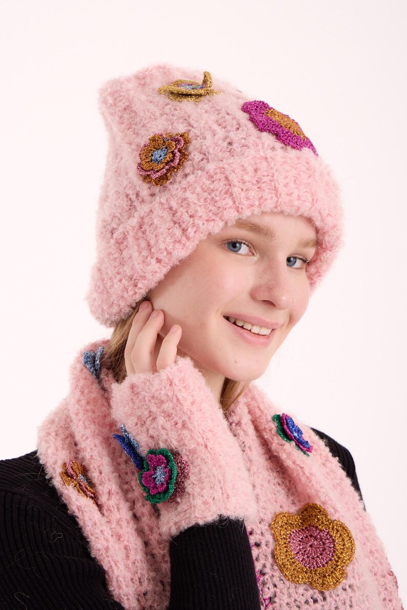 May include: A light pink knitted beanie and matching scarf set featuring a textured, fuzzy yarn construction. The set is decorated with multiple small, colorful crocheted flower appliqu&eacute;s in shades of gold, blue, purple, and magenta.