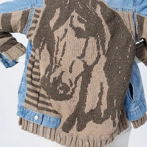 May include: A light blue denim jacket with a unique design. The back features a large, knitted horse head in shades of brown and tan. The sleeves have a striped pattern, and the jacket has a ribbed hem.